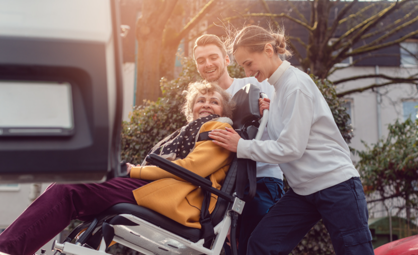 Older adult seated in a wheelchair being assisted by two caregivers beside a vehicle outdoors.